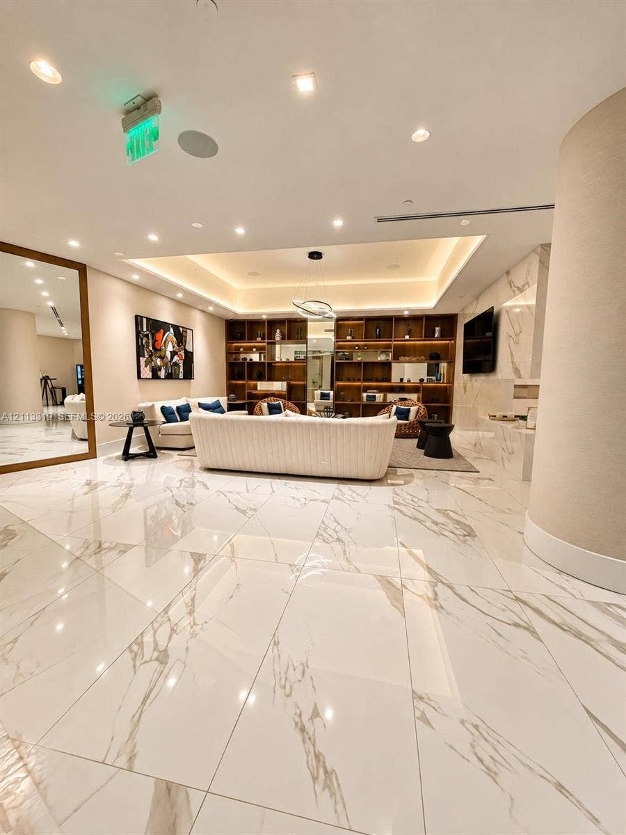 Large-format polished stone flooring, recessed tray ceiling with integrated cove lighting, floor-to-ceiling wooden built-in shelving, full-height wall mirror with metallic frame, and stone-surround fireplace feature