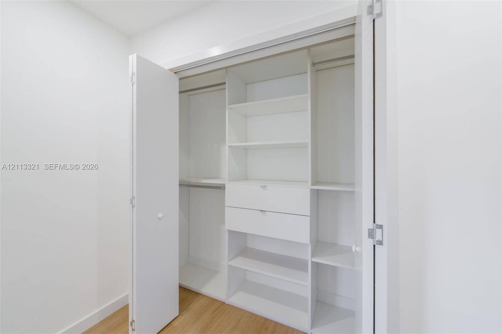 Custom reach-in closet featuring white built-in shelving, dual integrated drawers, hanging rods, and bi-fold doors, accented by wood-finish flooring