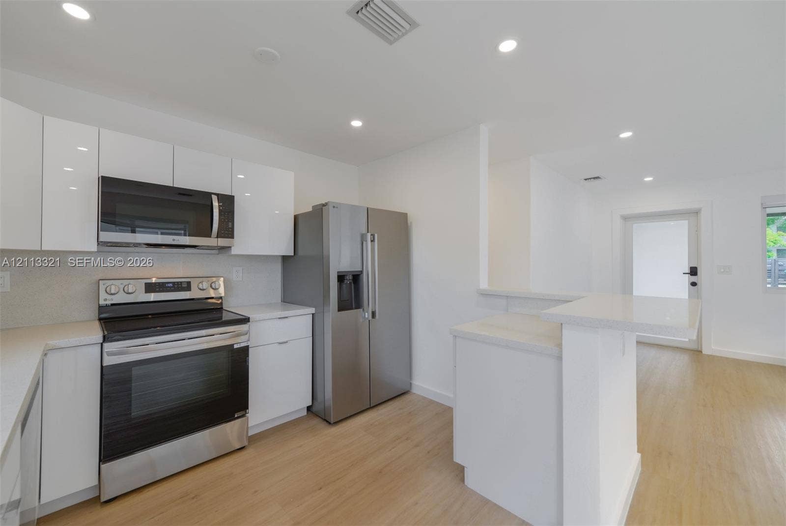 Modern kitchen featuring white flat-panel cabinetry, light-toned countertops, stainless steel appliances, wood-finish flooring, and recessed lighting