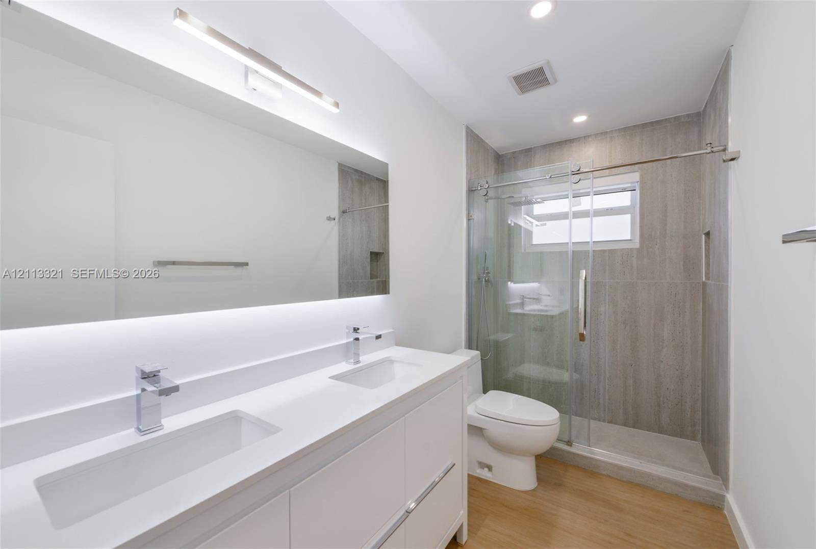 Dual-sink vanity with white stone-finish countertop, under-mount sinks, chrome faucets, full-width mirror with integrated lighting, and glass-enclosed shower with floor-to-ceiling tile