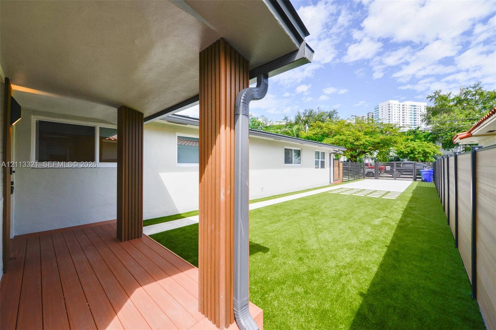 Covered patio with wood-finish decking and vertical slat support columns, featuring synthetic turf, concrete paver accents, and perimeter privacy fencing