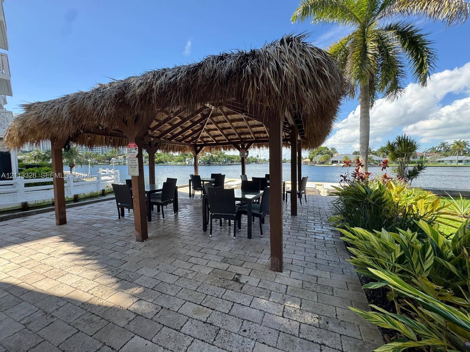 Hallandale Beach, FL 33009 Waterfront gazebo with a thatched roof, supported by timber posts, situated on a stone paver patio