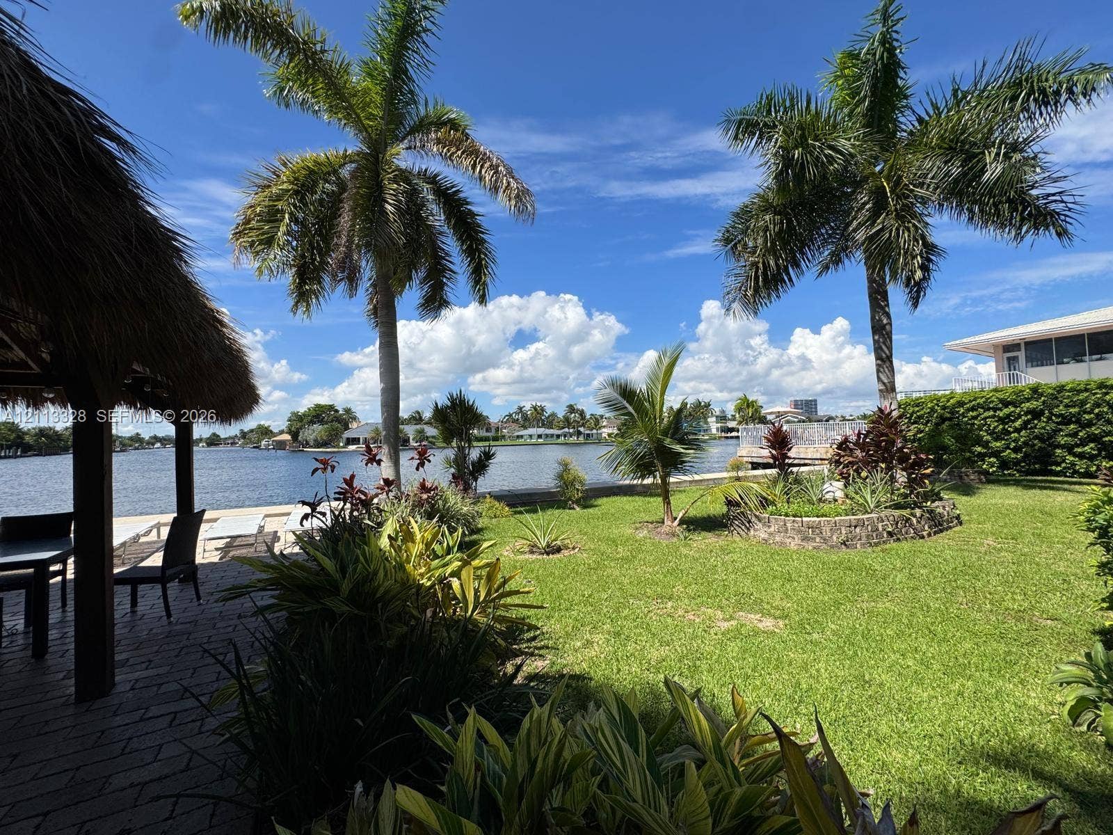 Hallandale Beach, FL 33009 Waterfront backyard featuring a thatched-roof tiki hut, paved patio, expansive lawn, mature palm trees, and a concrete seawall