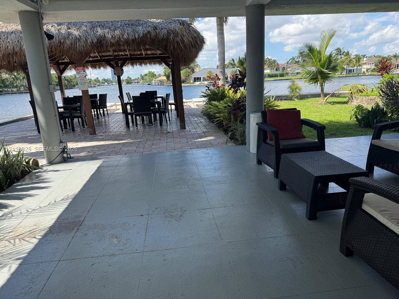 Hallandale Beach, FL 33009 Covered patio with tile flooring and structural columns, featuring a waterfront paver area, a thatched-roof tiki hut, and mature landscaping