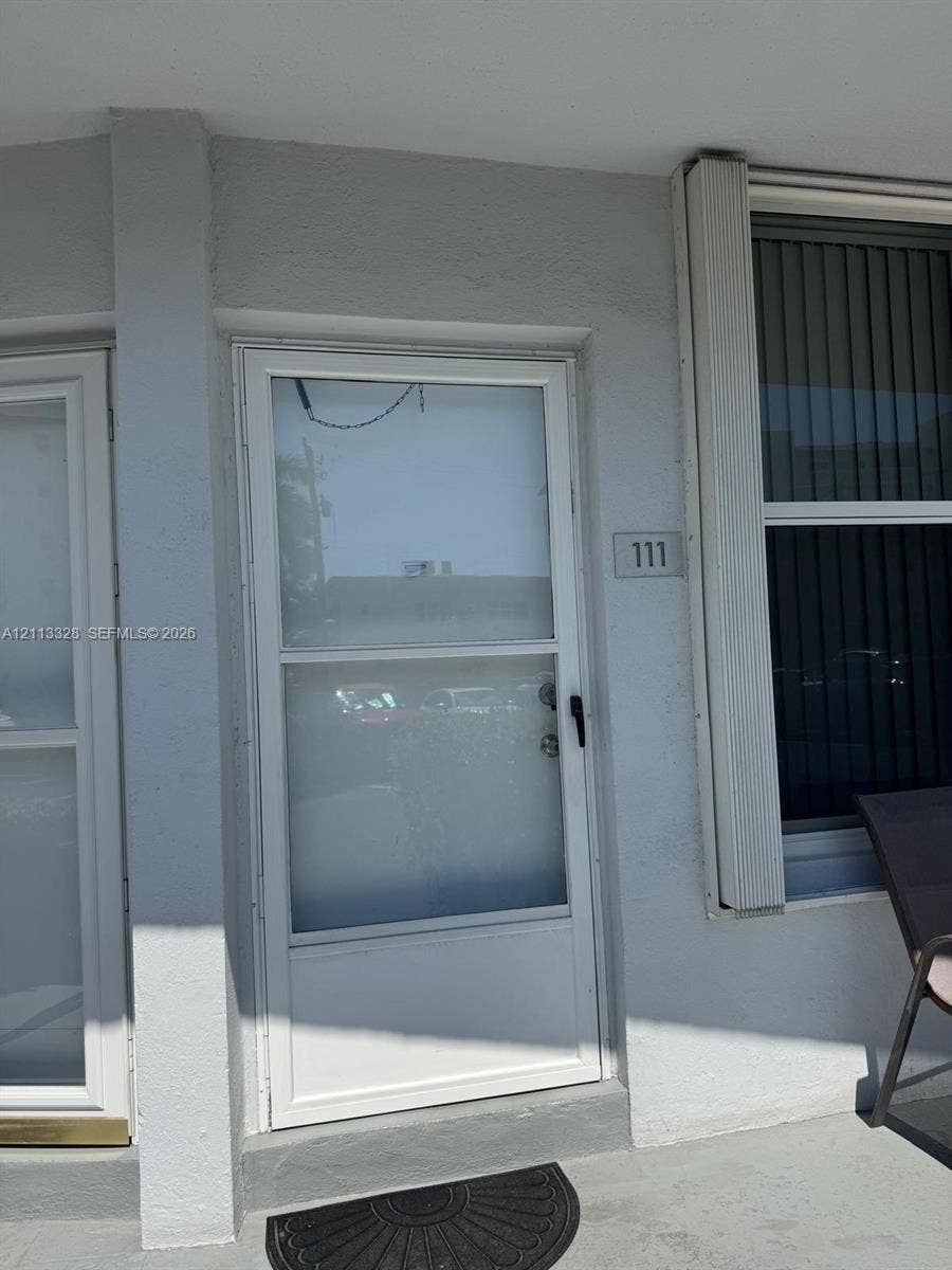 Hallandale Beach, FL 33009 Exterior entry featuring a glass-panel storm door, light-colored smooth stucco siding, vertical window casing, and a dedicated address plaque