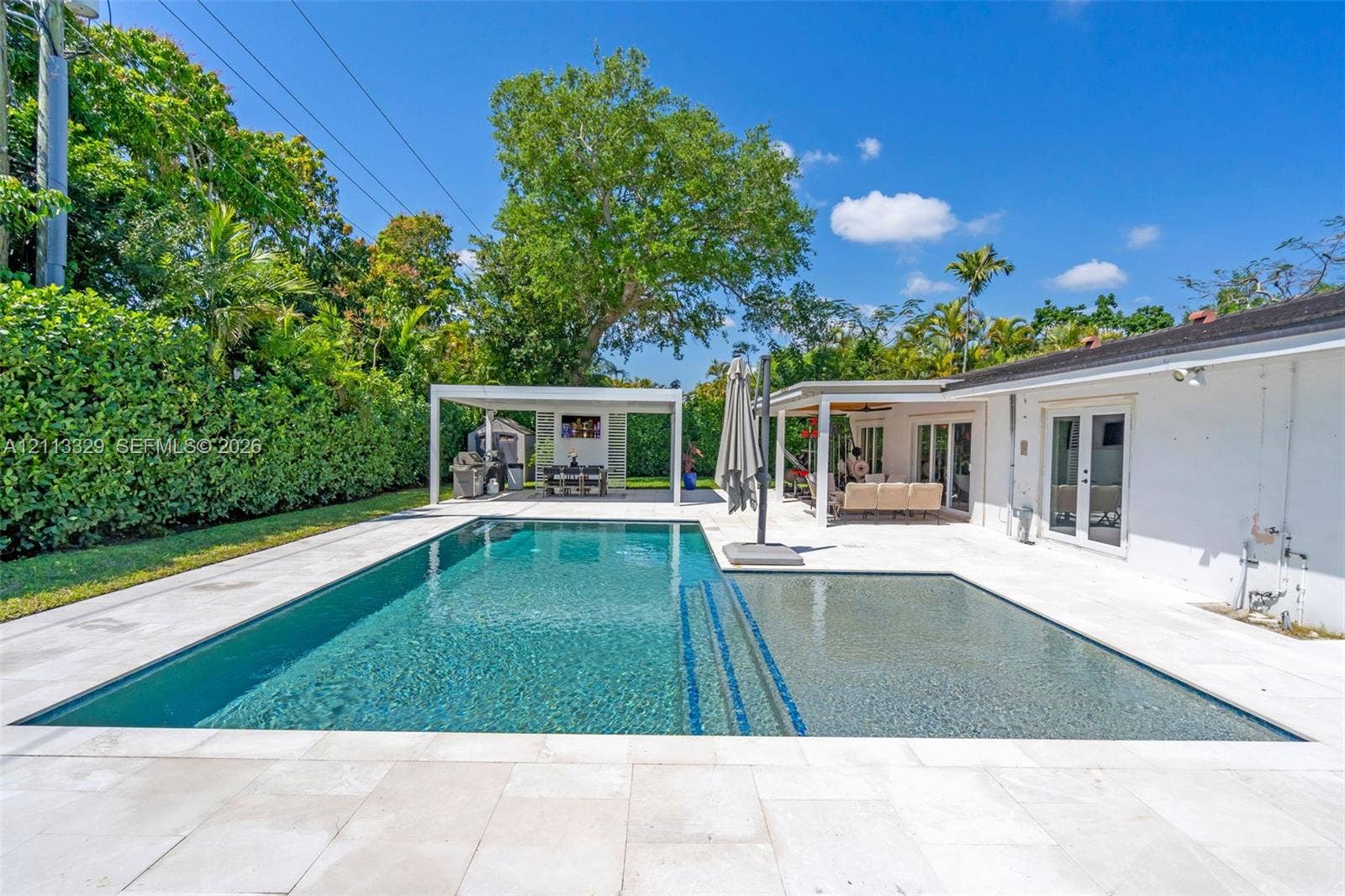 6440 SW 144th Street, Coral Gables, FL 33158 Swimming pool with a sun shelf and built-in steps, surrounded by expansive light-toned stone patio pavers, featuring a detached covered pavilion and a covered exterior porch