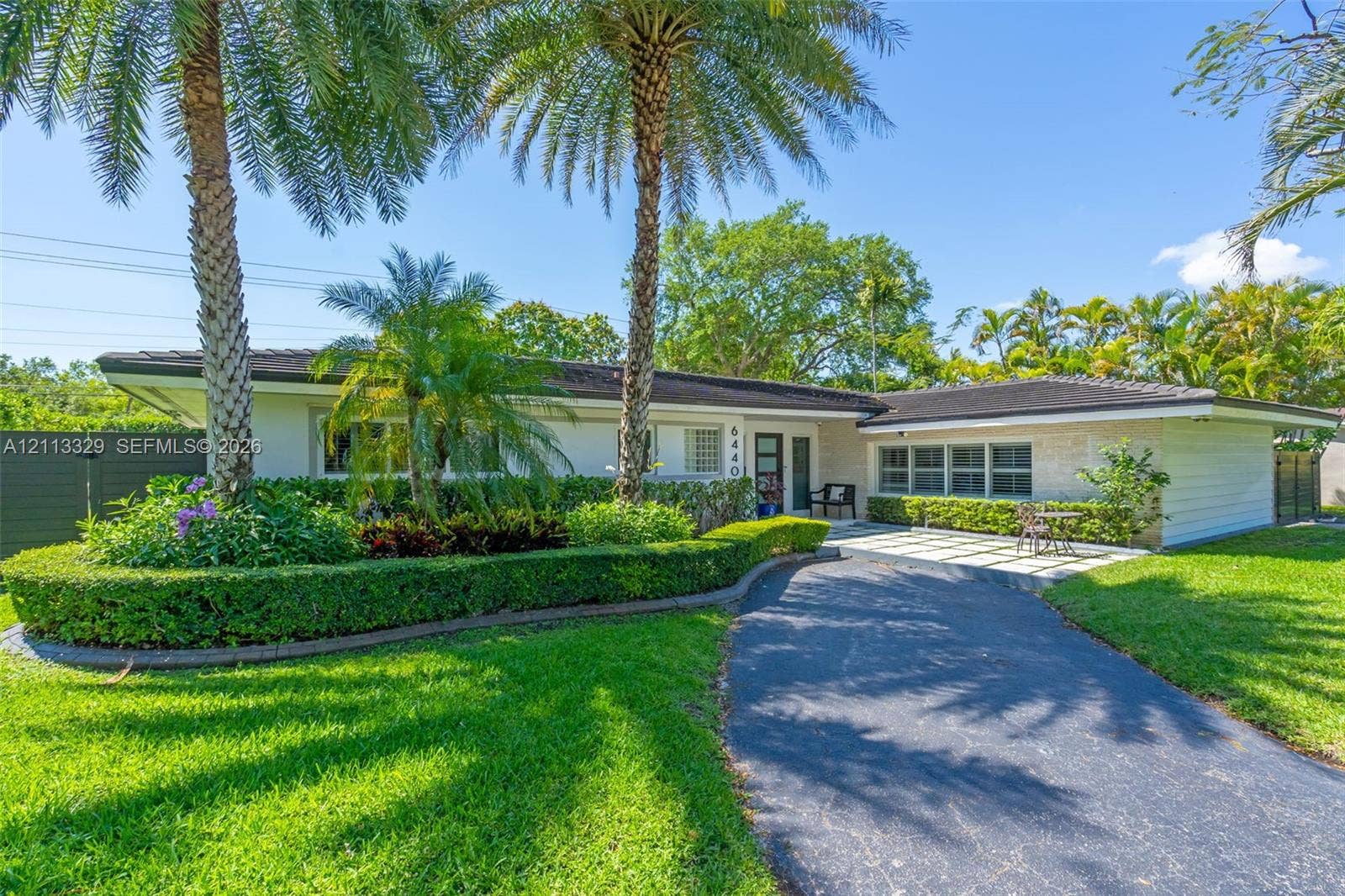 6440 SW 144th Street, Coral Gables, FL 33158 Single-story residence featuring a low-pitched roof, manicured hedges, mature palm trees, stone-finish exterior accents, and a paved front patio area