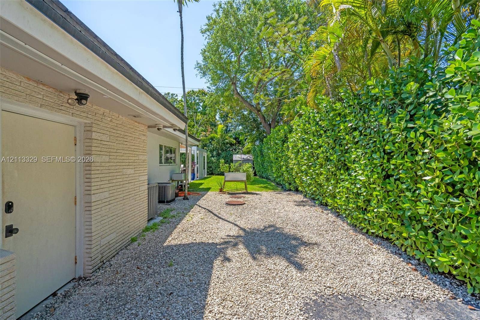 6440 SW 144th Street, Coral Gables, FL 33158 Side exterior featuring painted brick siding, gravel ground cover, and a privacy hedge of mature greenery