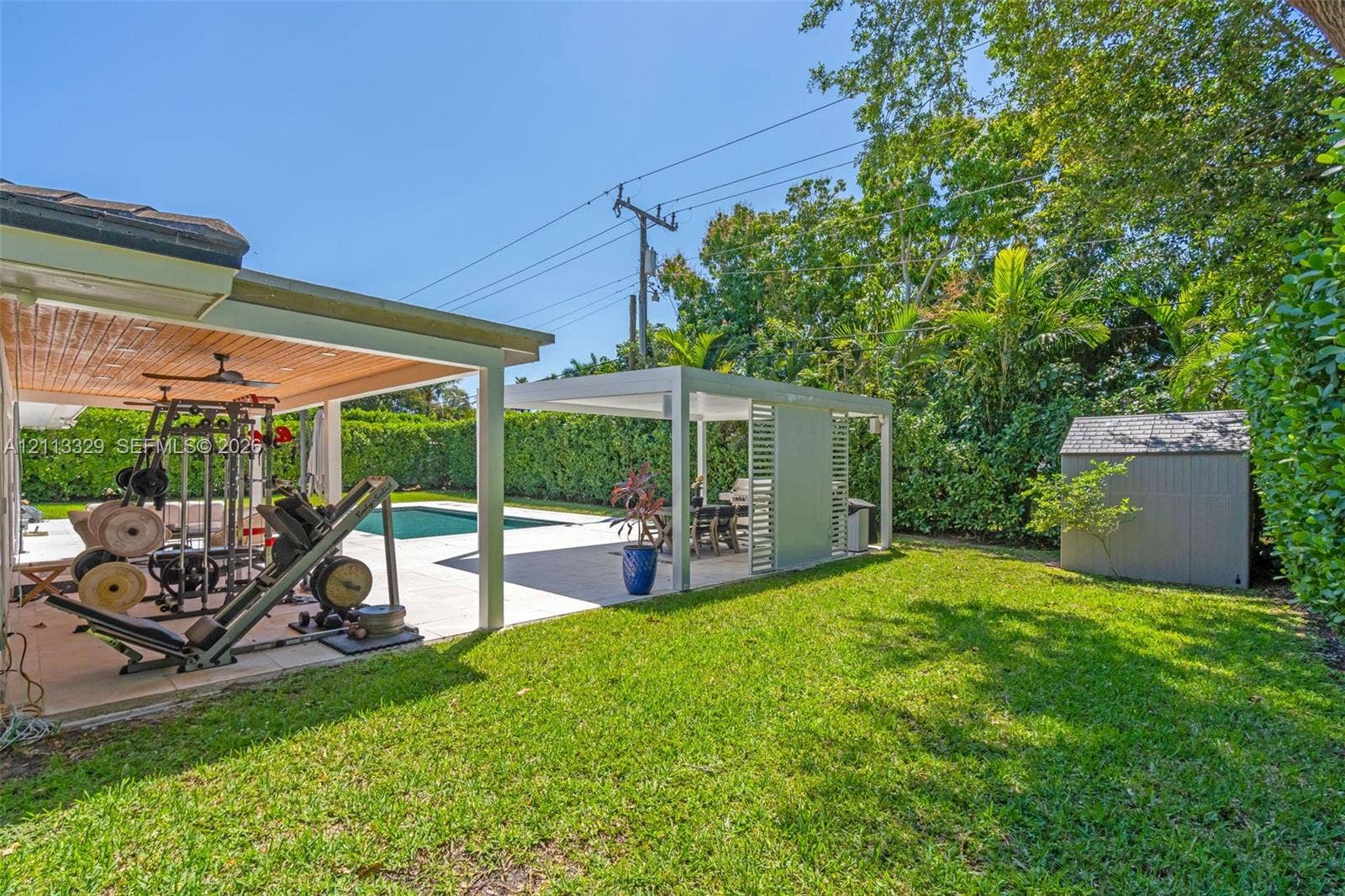 6440 SW 144th Street, Coral Gables, FL 33158 Backyard featuring a swimming pool, a covered patio with a beadboard ceiling, a separate metal-framed pergola, and a storage shed surrounded by mature perimeter hedging