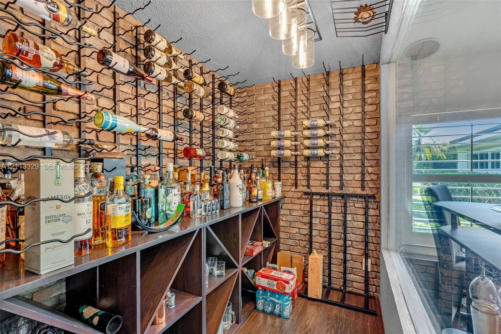 6440 SW 144th Street, Coral Gables, FL 33158 Custom beverage storage space featuring a full-wall brick accent, integrated floor-to-ceiling metal racking, built-in wooden shelving with geometric cubbies, and overhead glass pendant lighting