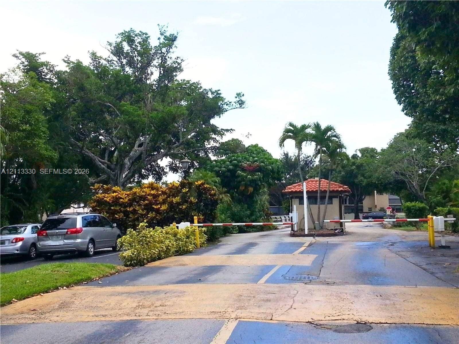 6001 N Falls Cir Drive, #105, Lauderhill, FL 33319 Gated entry featuring a controlled-access barrier arm, guard house with a terracotta tile roof, and mature tropical landscaping