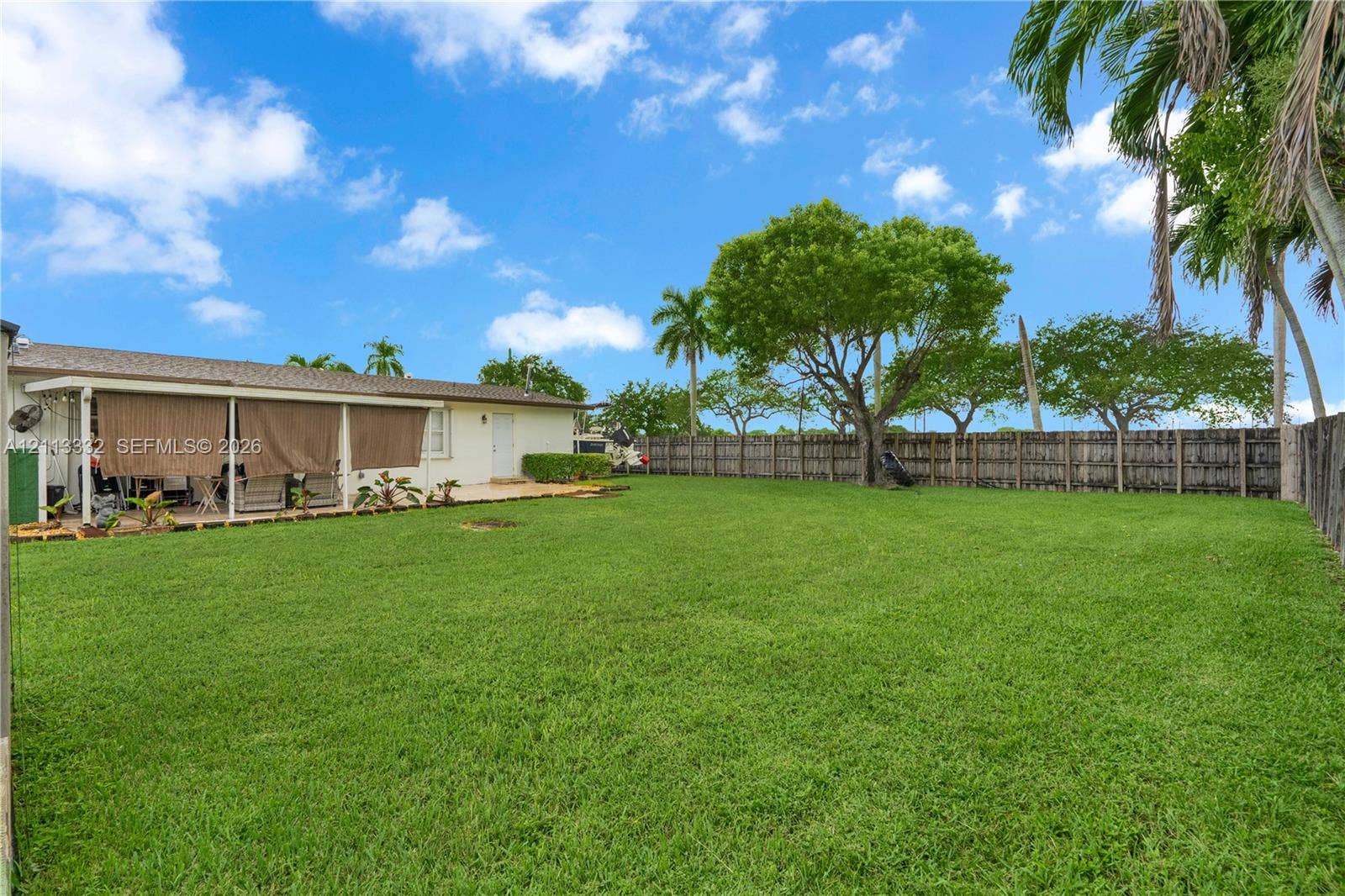 13111 SW 84th Street, Miami, FL 33183 Expansive backyard featuring a large grass lawn, perimeter wood fencing, mature trees, and a covered patio area