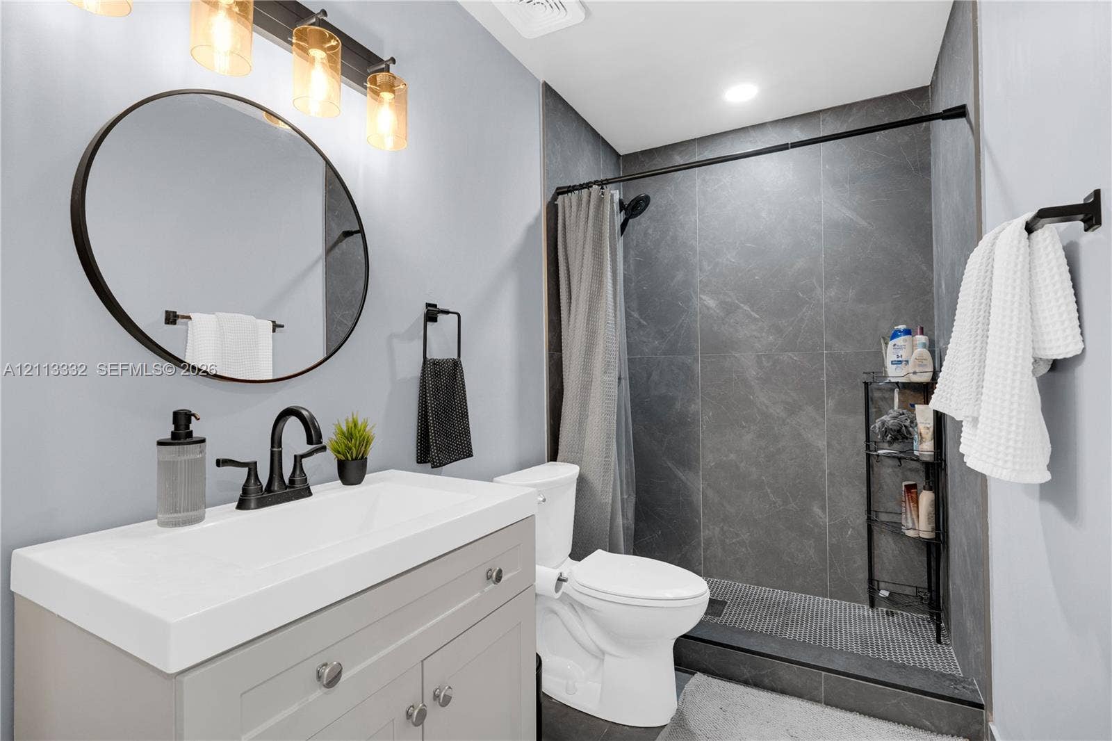 13111 SW 84th Street, Miami, FL 33183 Bathroom featuring a vanity with a rectangular sink, matte black hardware, a round mirror, and a walk-in shower with dark tile surround