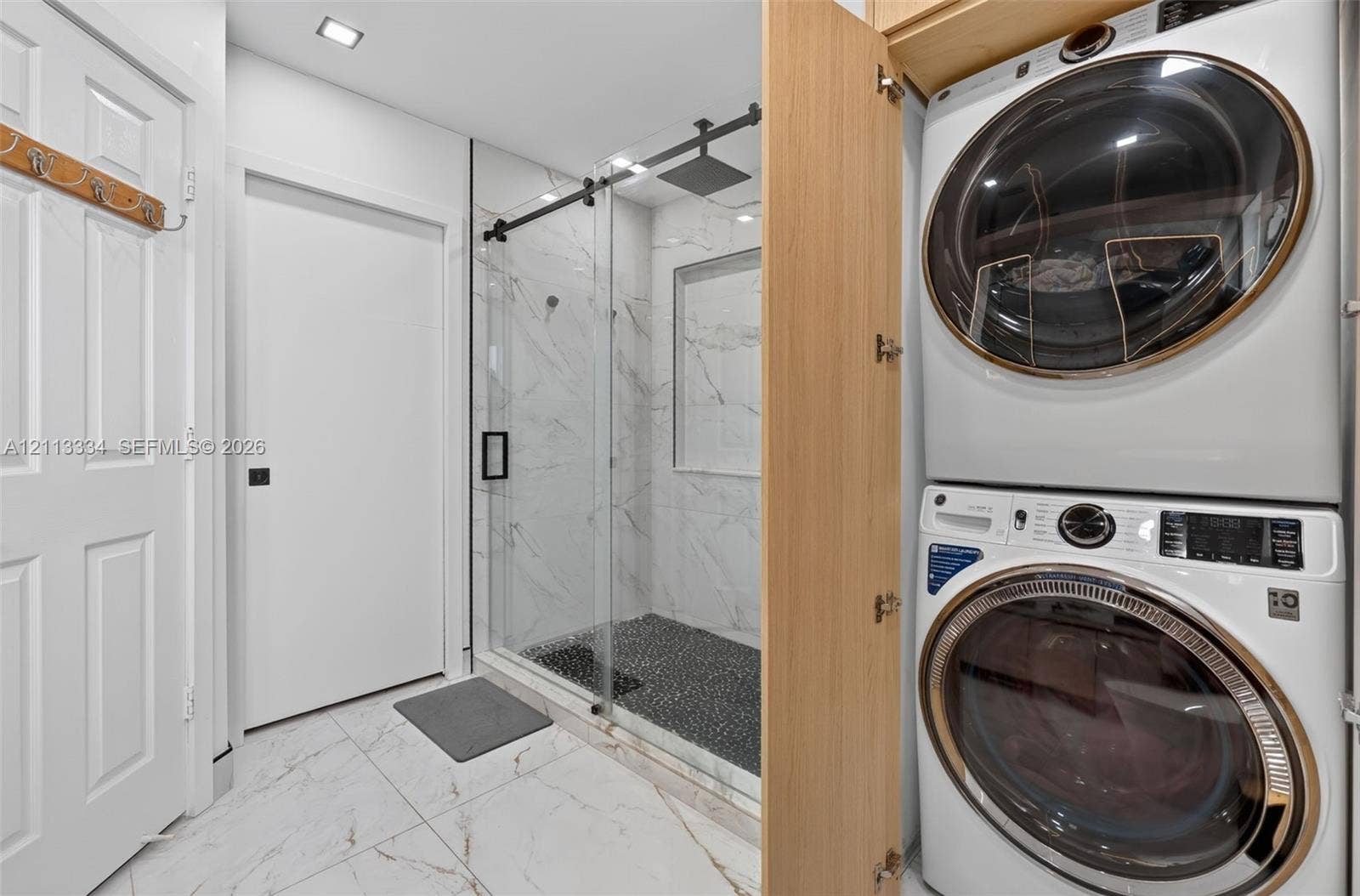 28232 SW 128th Path, Homestead, FL 33033 Walk-in shower with stone-look wall tile, recessed shelving, a glass sliding door with matte black hardware, and a pebble-tiled floor, featuring built-in stacked laundry appliances behind cabinetry