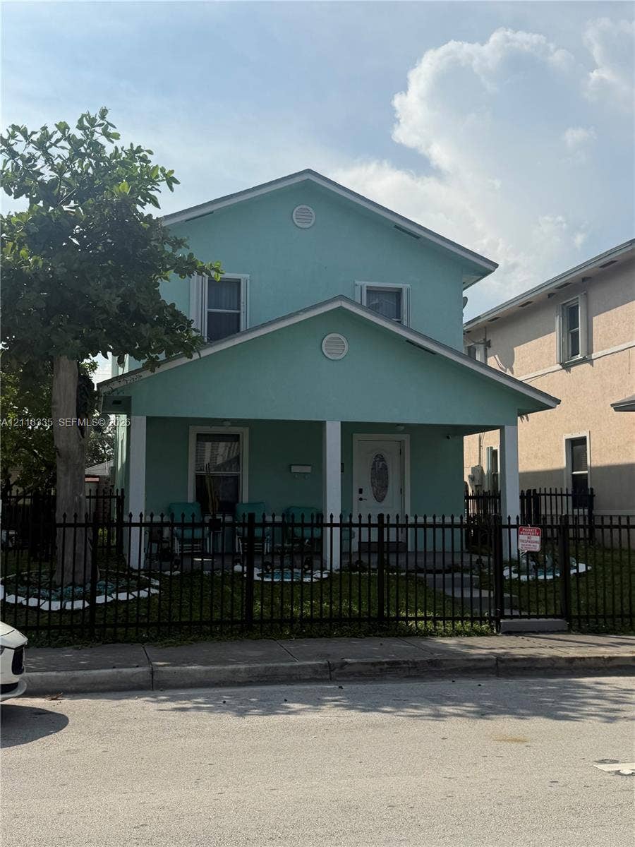 1990 NW 1st Place, Miami, FL 33136 Two-story residence featuring a covered front porch, white support columns, dual-gabled roofline, and metal perimeter fencing