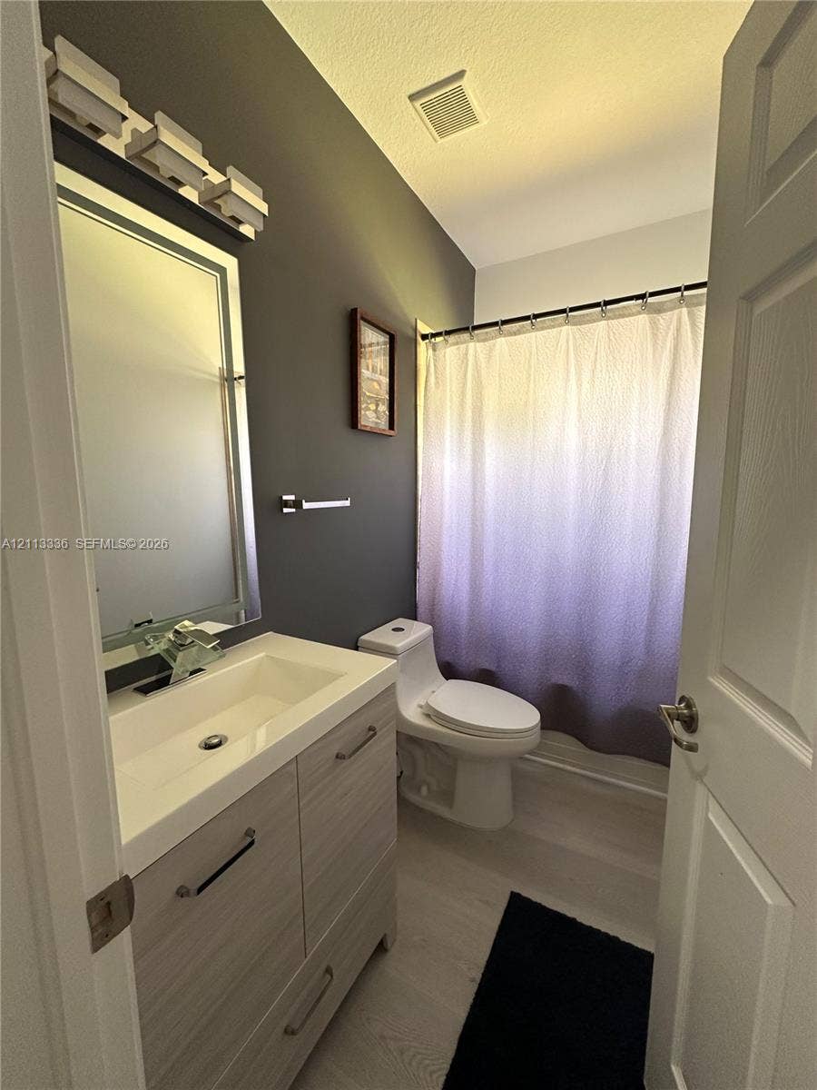 Bathroom featuring a wood-finish vanity with a rectangular sink, integrated mirror with light fixture, high-profile toilet, and wood-finish flooring