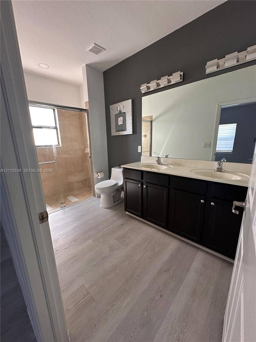 Dual-sink vanity with dark cabinetry and light-toned countertop, large wall-mounted mirror, two multi-bulb vanity light fixtures, wood-finish flooring, and a walk-in shower with a glass enclosure