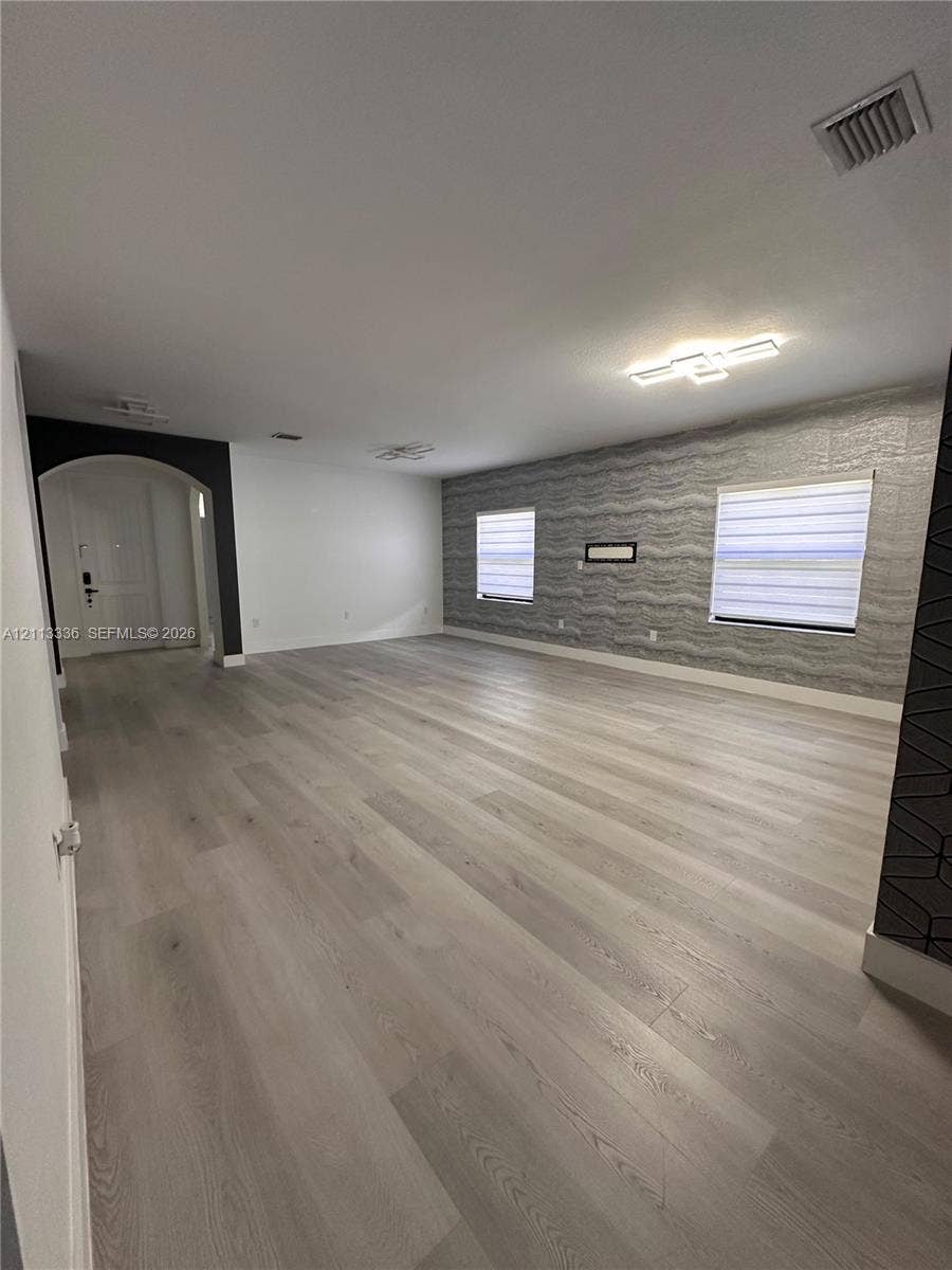Open interior space featuring light-toned wood-finish flooring, a textured accent wall, integrated flush-mount ceiling lighting, and a prominent arched entryway