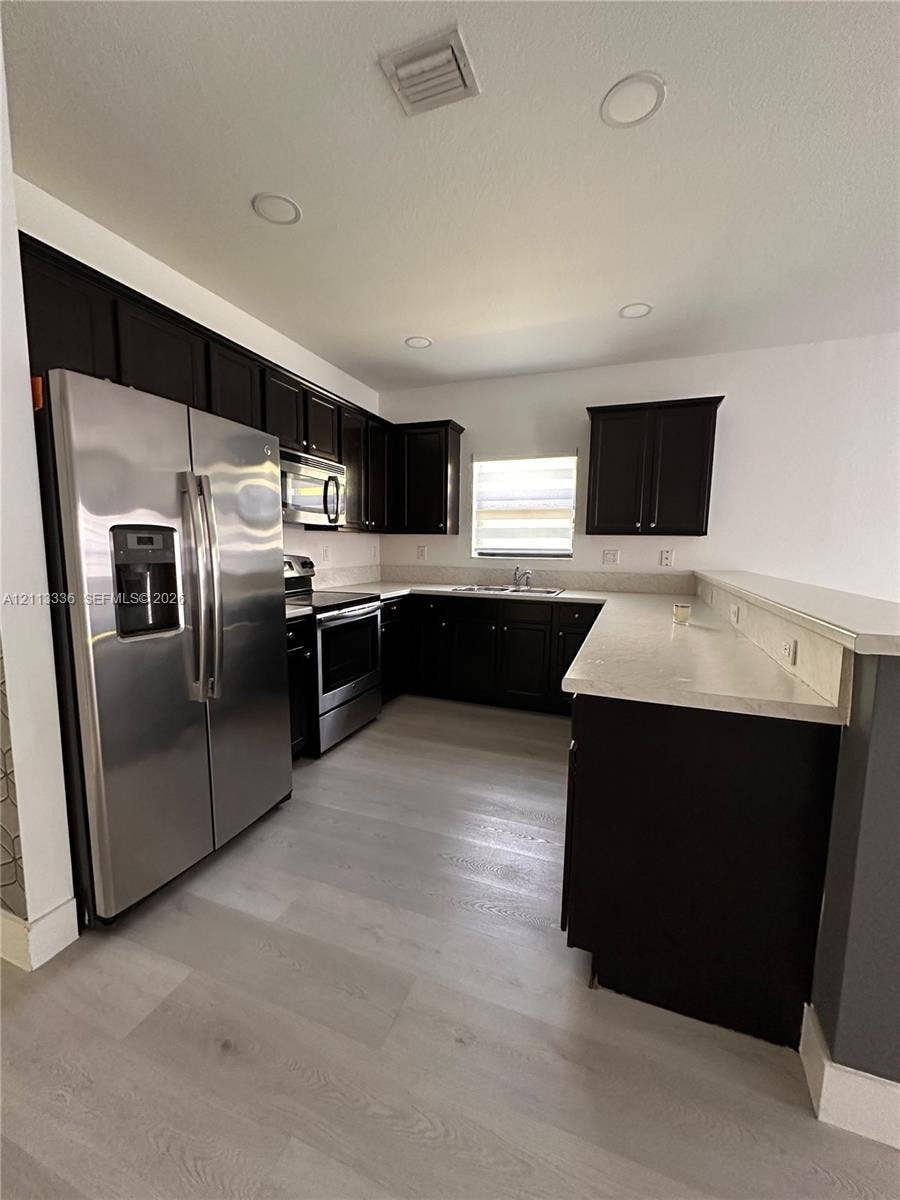 Kitchen featuring dark-finish cabinetry, light-toned stone-style countertops, stainless steel appliances, and wood-finish flooring
