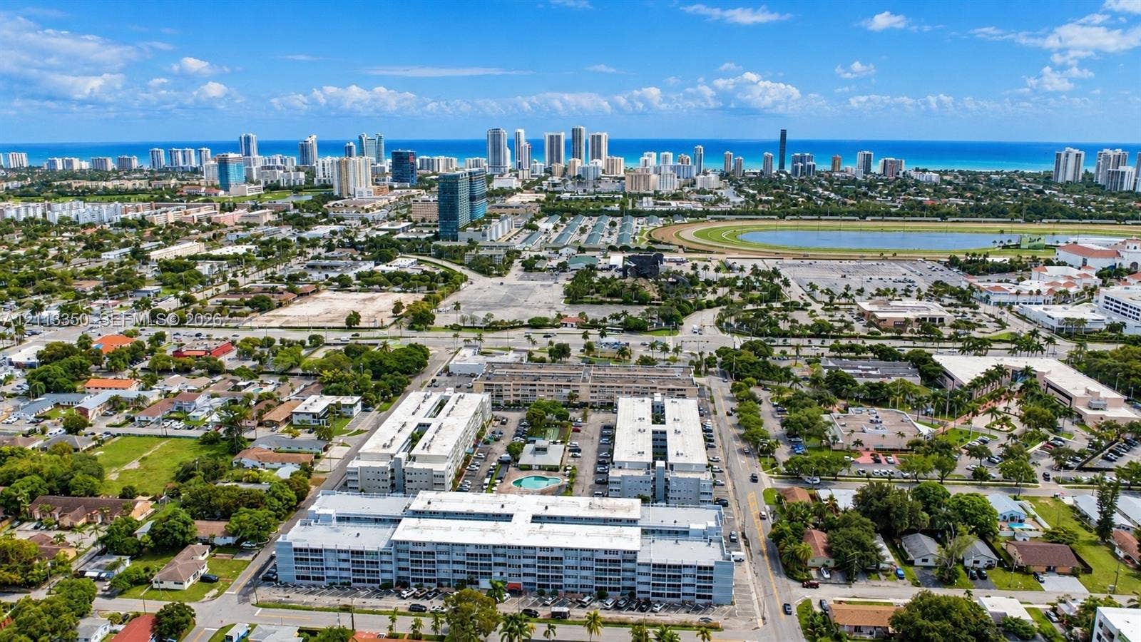 Aerial perspective of a multi-story residential complex featuring a central outdoor pool, landscaped grounds, and proximity to the coastline and metropolitan skyline