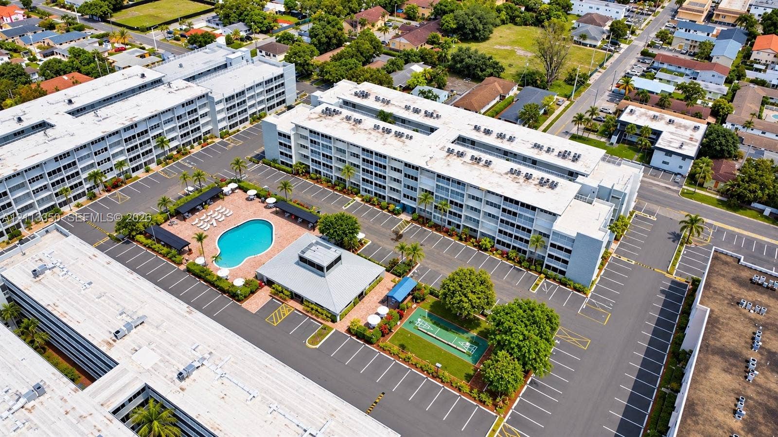 Multi-story residential complex featuring an outdoor swimming pool, paved surface parking, a clubhouse structure, and an on-site shuffleboard court