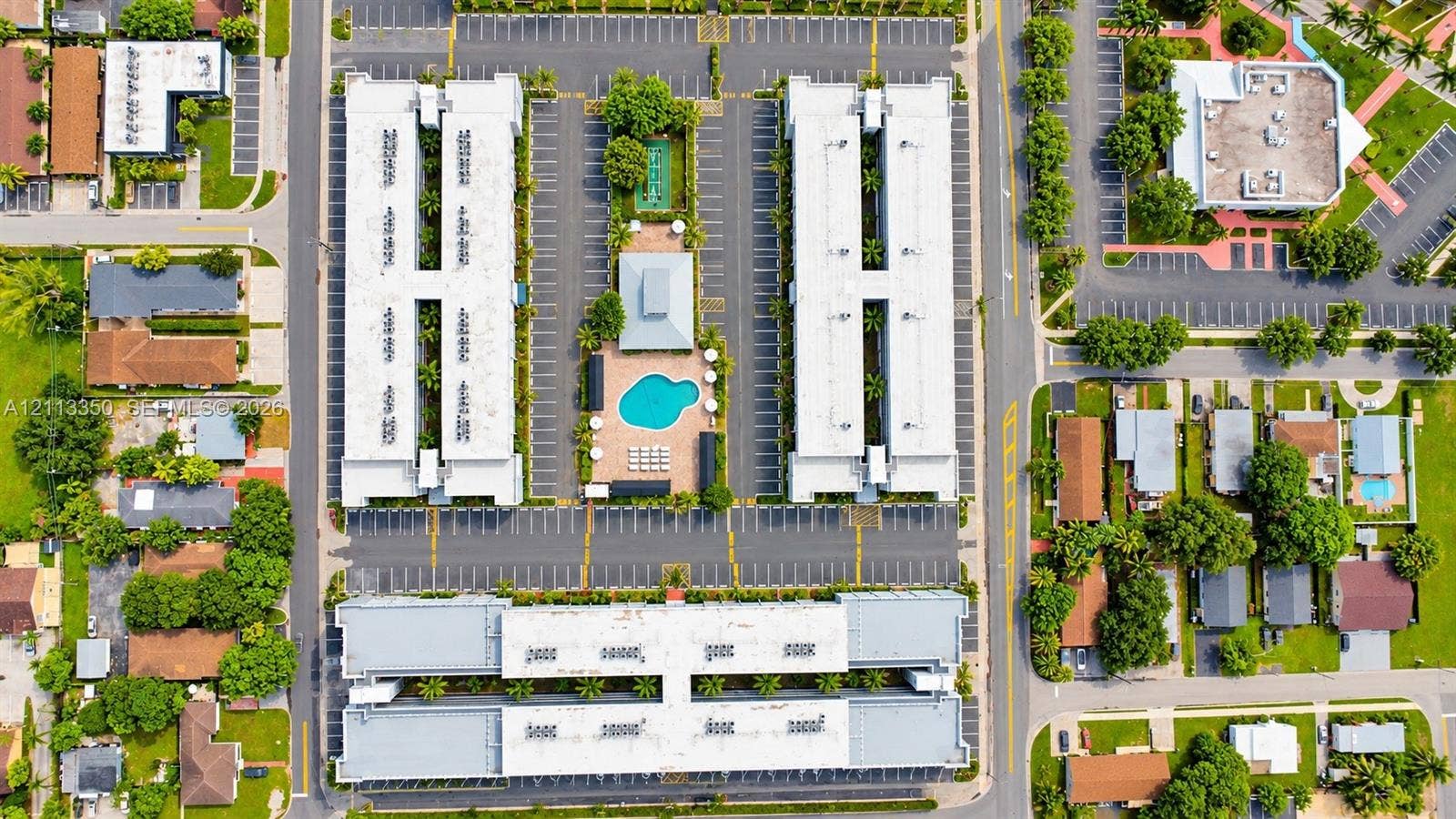 Aerial perspective of a multi-building residential complex featuring a central outdoor swimming pool, paved surface parking, paved walking paths, and mature landscaping