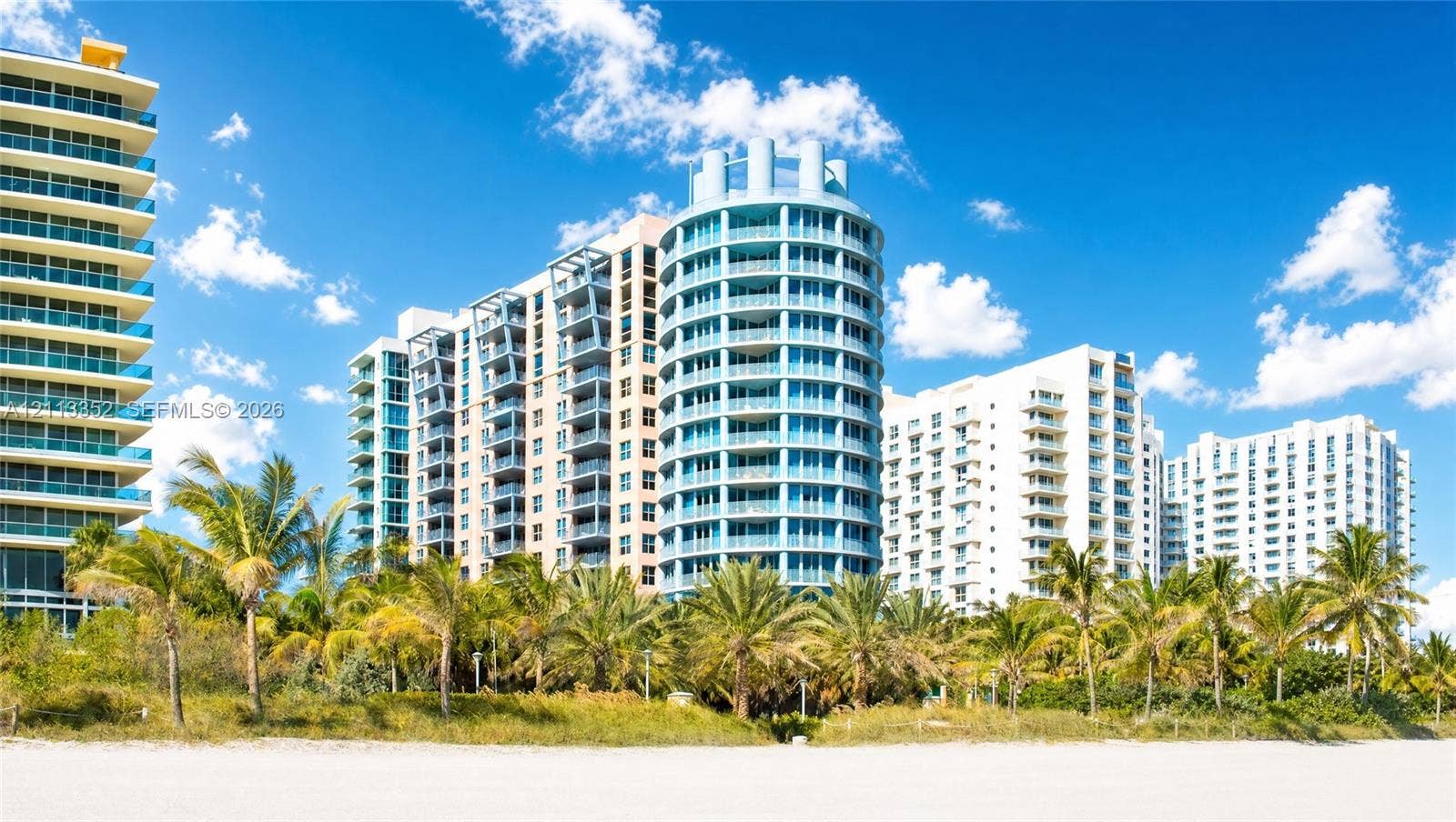 Multi-story high-rise architecture featuring expansive glass-paneled balconies, cylindrical structural elements, and a white sand beach perimeter