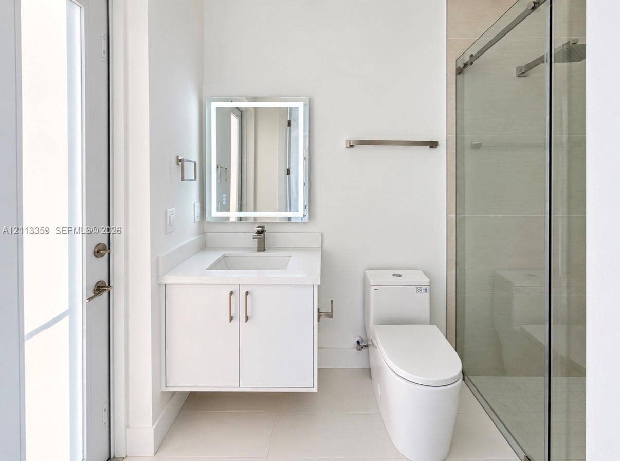 Floating vanity featuring a solid surface countertop, integrated sink, and brushed metal faucet, complemented by an illuminated rectangular mirror, a one-piece toilet, and a glass-enclosed shower with tile surround