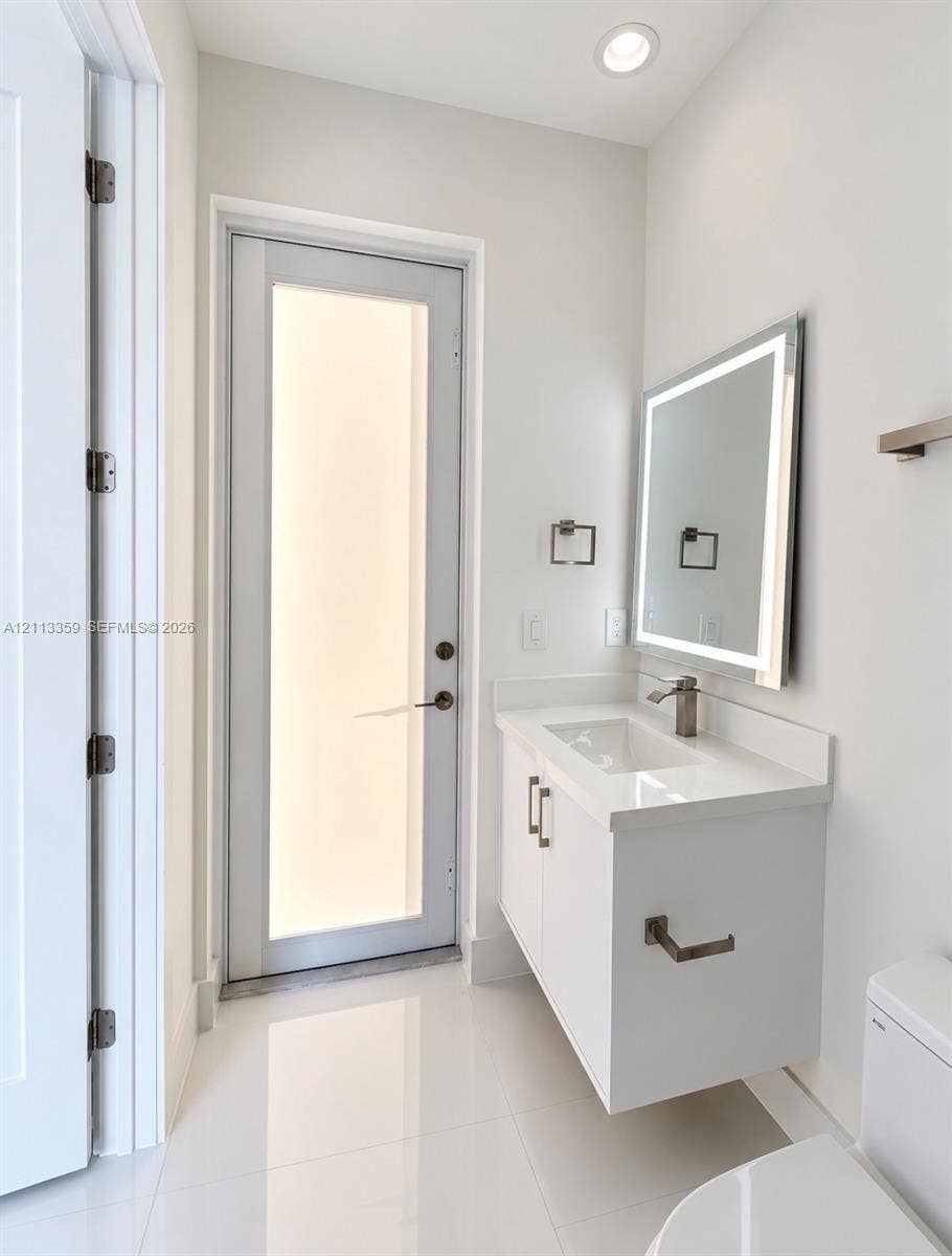 Floating vanity with white cabinetry, rectangular basin, integrated countertop, and brushed metal hardware, complemented by a backlit rectangular mirror, polished tile flooring, and a glass-panel exterior door