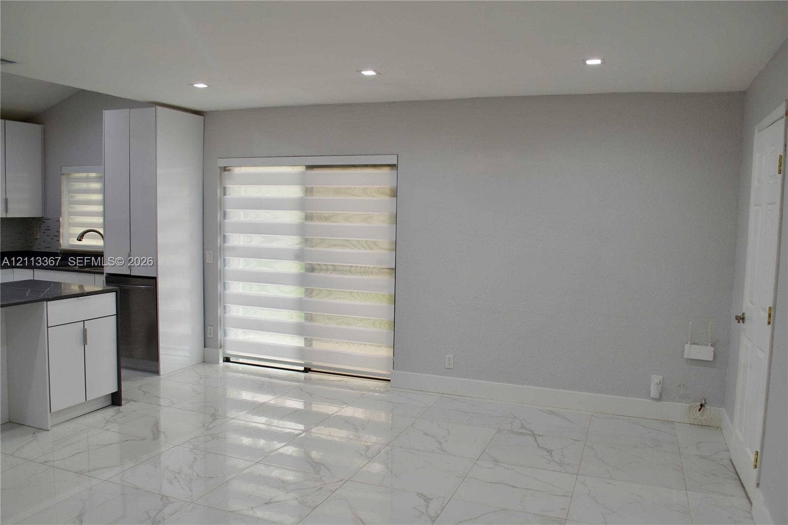 Large-format tile flooring with marble-patterned veining, recessed lighting, white flat-panel cabinetry, dark stone countertops, and dual-layered sliding glass door blinds
