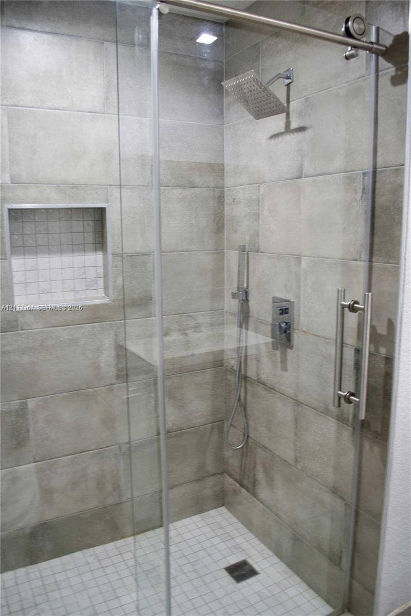 Walk-in shower featuring large-format neutral tile walls, a sliding glass door with stainless steel hardware, a rainfall showerhead, a handheld sprayer, and a built-in recessed niche