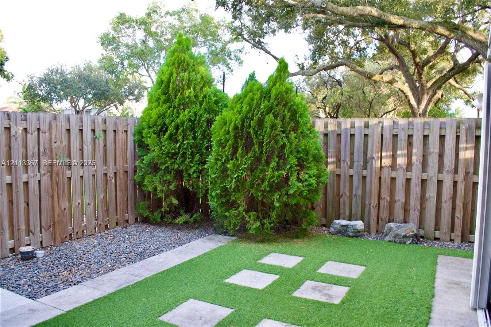 Fenced outdoor space featuring synthetic turf, concrete stepping stones, stone ground cover, and mature evergreen privacy plantings
