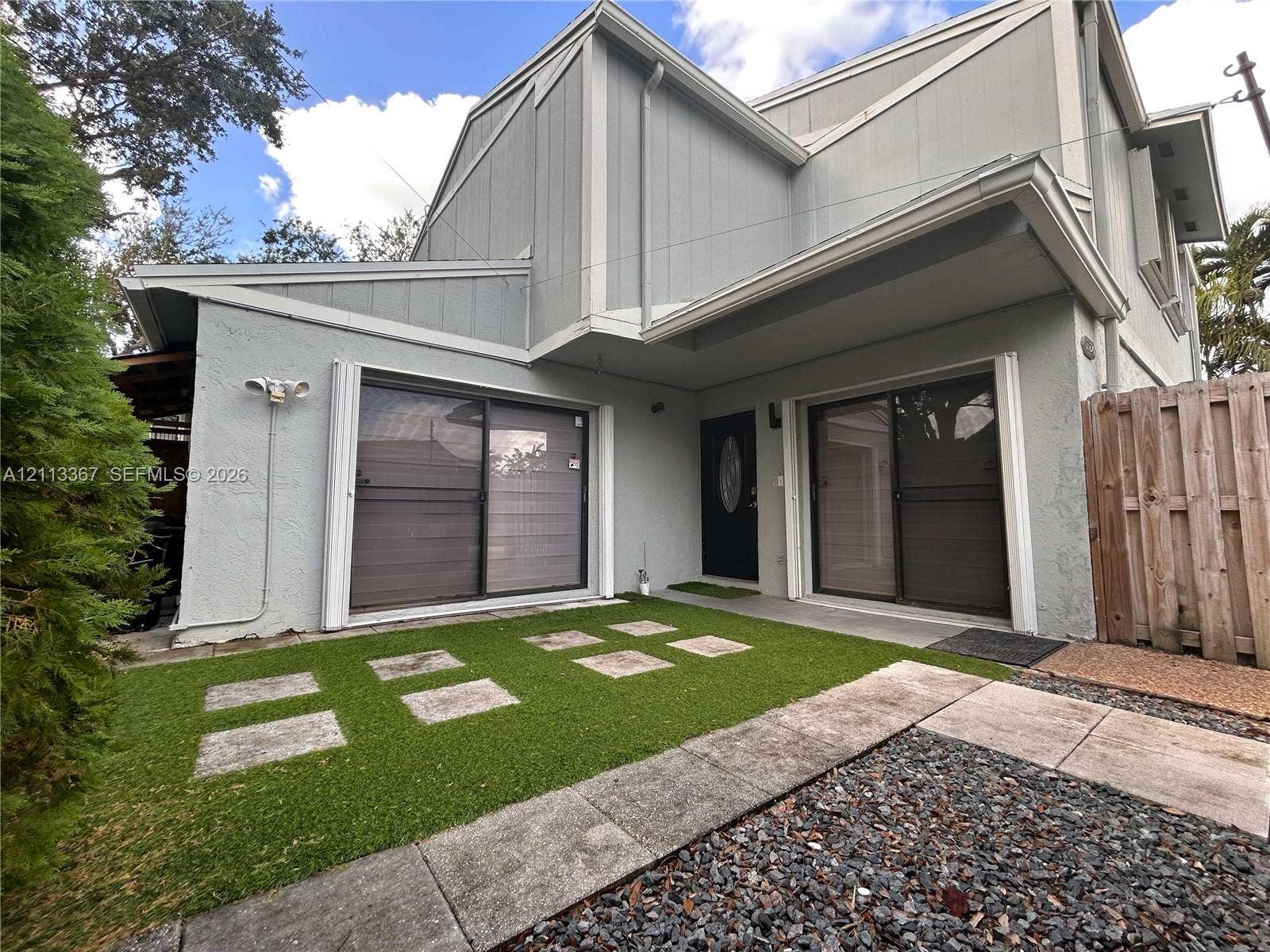 Multi-level exterior featuring light-toned siding, dual sliding glass doors, a synthetic turf yard with stone paver inlays, and a vertical wood privacy fence