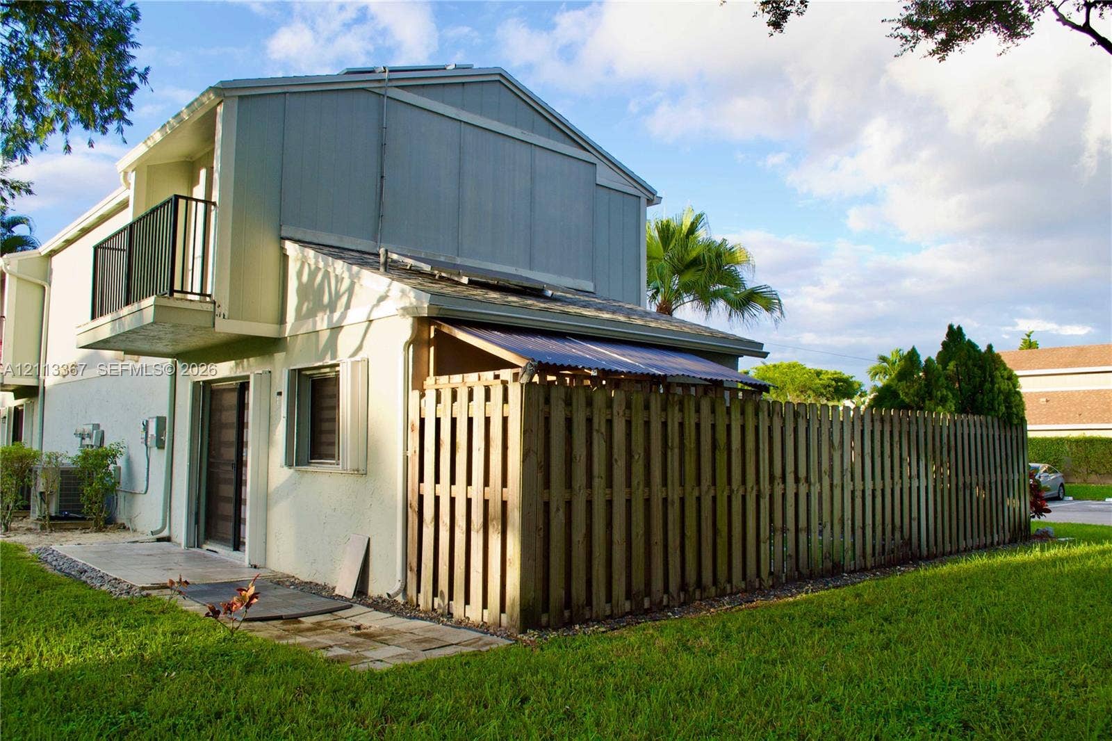 Two-story exterior featuring light-toned vertical siding, a private balcony with metal railing, a covered patio area, and an enclosed wooden privacy fence
