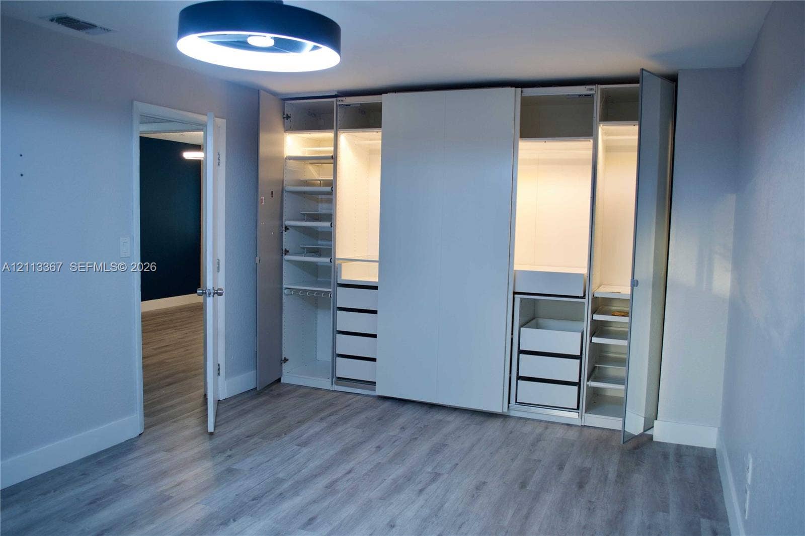 Custom floor-to-ceiling built-in wardrobe system with integrated drawer storage, adjustable shelving, and interior lighting, complemented by wood-finish flooring and a contemporary circular flush-mount ceiling fixture