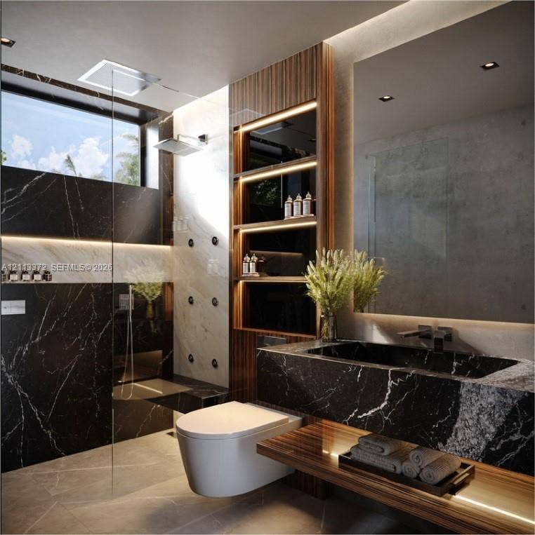 3312 NE 16th Court, Fort Lauderdale, FL 33305 Black stone vanity with an integrated basin, wall-mounted commode, glass-enclosed shower featuring body sprays, wood-finish built-in shelving with integrated lighting, and a large rectangular mirror with perimeter illumination