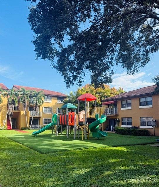 15610 SW 80th Street, #J-103, Miami, FL 33193 Outdoor recreation area featuring a multi-component play structure with slides and climbing equipment, situated on a manicured lawn with mature trees