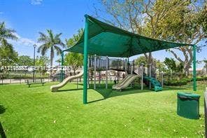Outdoor recreational area featuring a playground structure with dual slides, protective shade canopy, synthetic turf surfacing, and surrounding mature trees