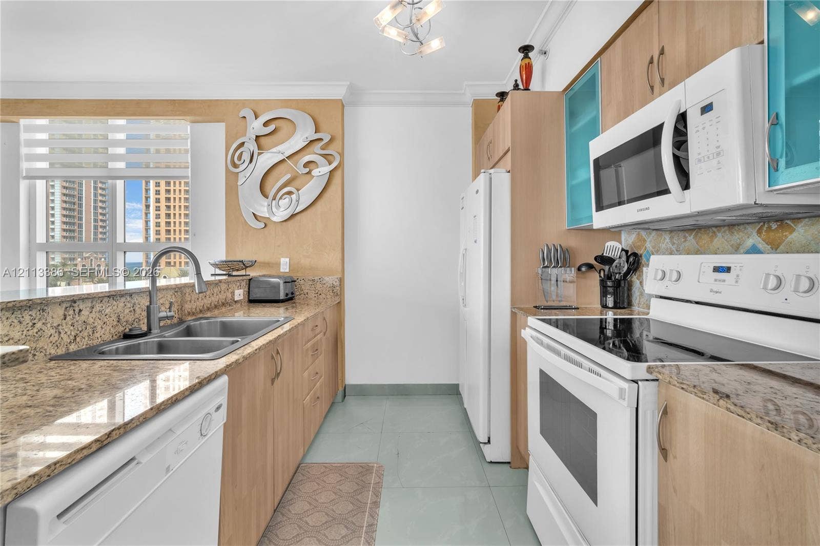 Kitchen featuring light wood-finish cabinetry, stone-surface countertops, dual-basin stainless steel sink, tile flooring, and integrated white appliances