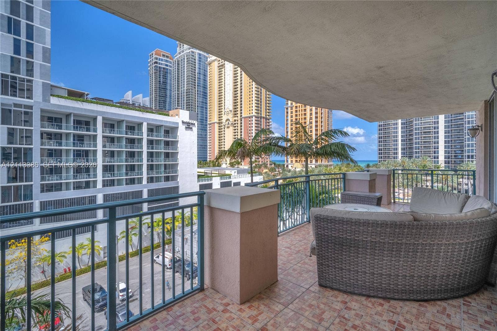 Covered balcony featuring tile flooring and metal railings, overlooking urban surroundings with palm trees and ocean glimpses