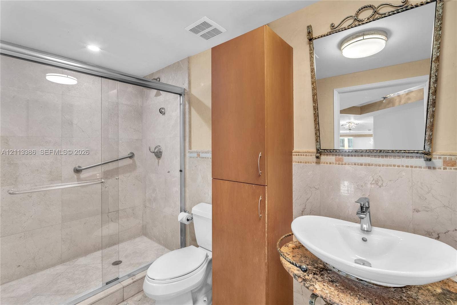 Walk-in shower with sliding glass enclosure and tiled walls, stone-finish countertop with vessel sink, tall built-in wooden cabinetry, and decorative wall mirror