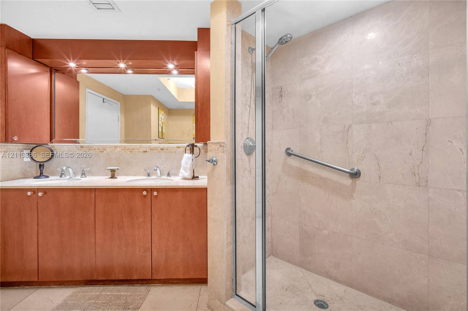 Dual-sink vanity with light stone countertop and wood-finish cabinetry, frameless glass shower enclosure, and neutral-toned stone wall tile