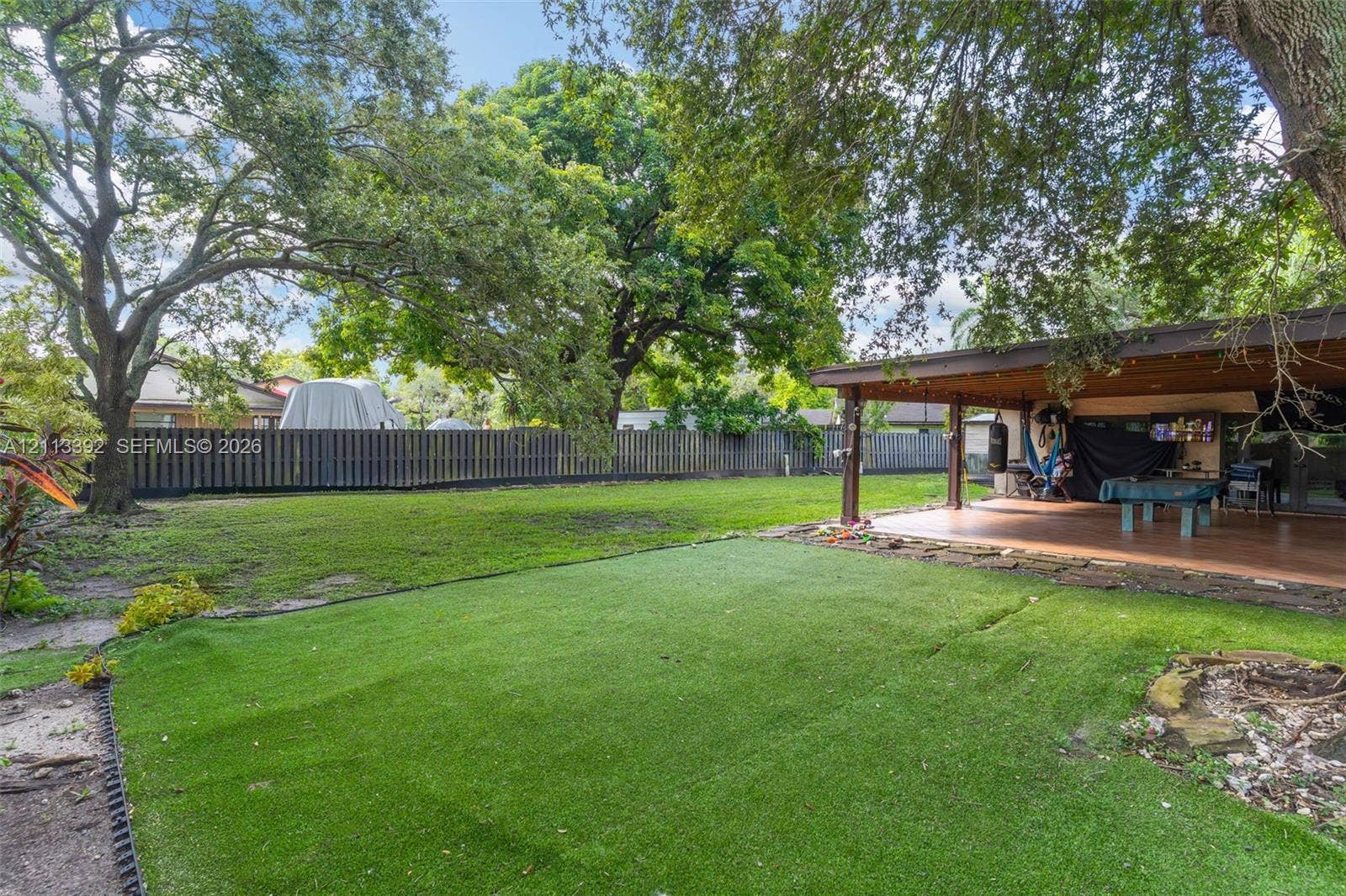 10705 SW 134th Court, Miami, FL 33186 Expansive backyard featuring a covered patio, synthetic turf lawn, perimeter wood fencing, and mature canopy trees