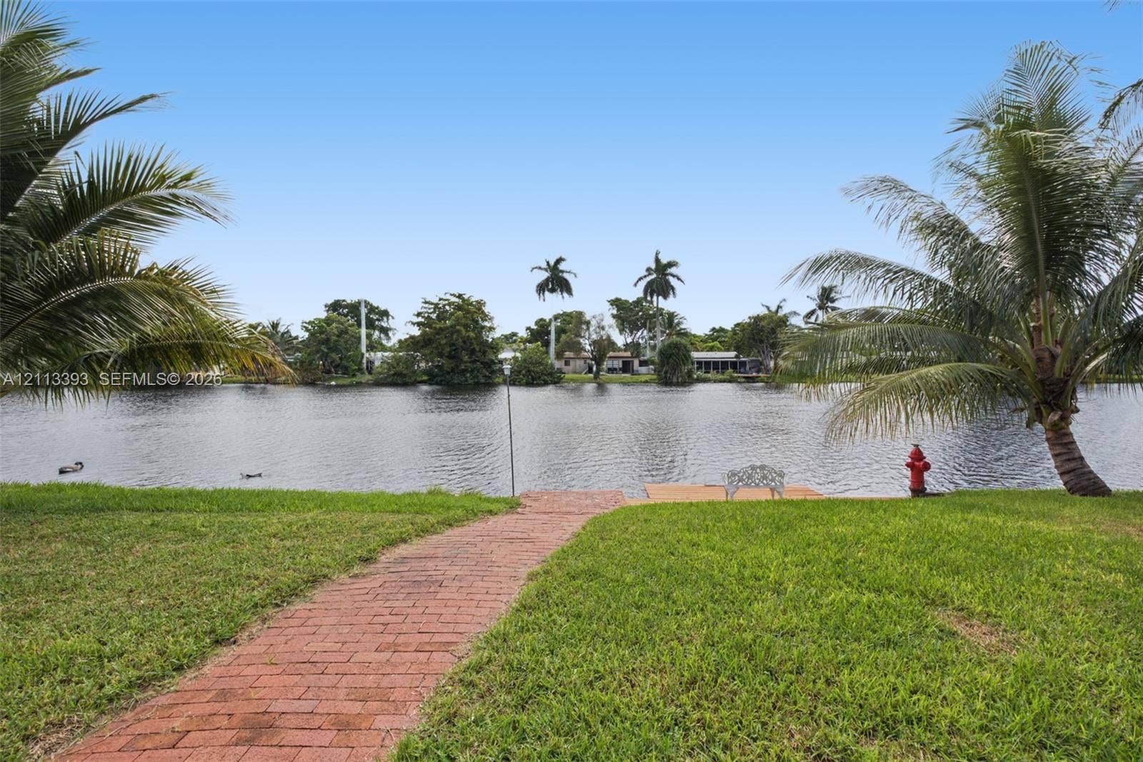 4136 SW 49th Street, Dania Beach, FL 33314 Waterfront parcel featuring a brick paver walkway, manicured lawn, and mature palm trees
