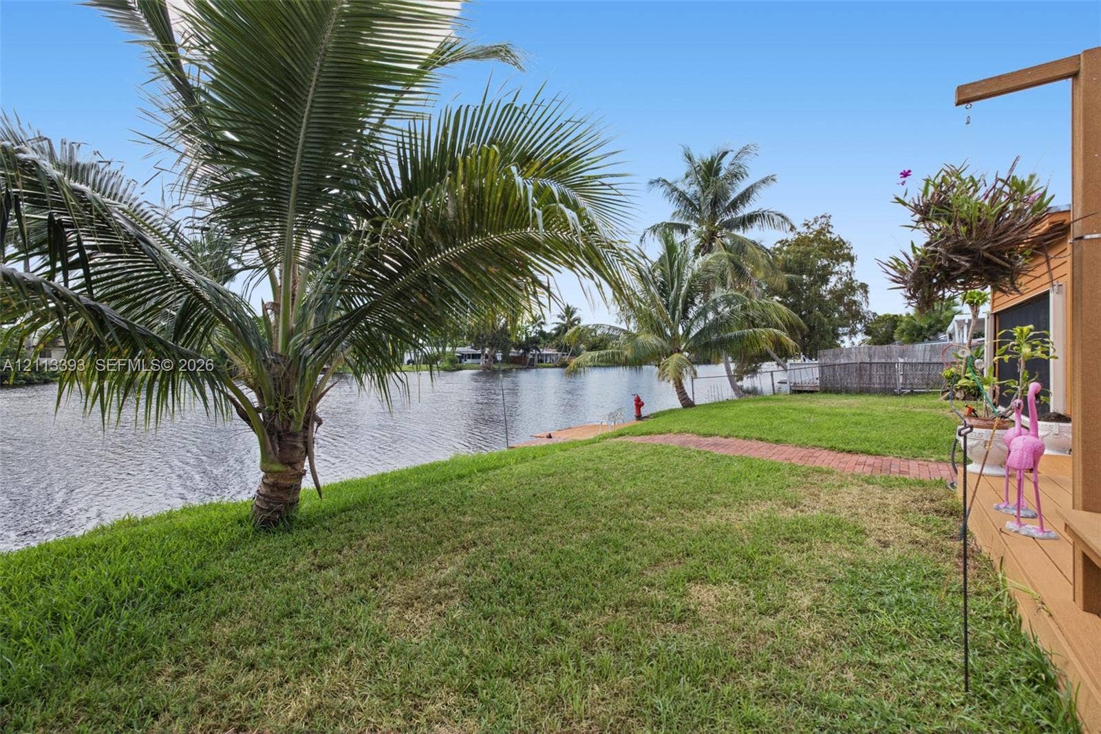 4136 SW 49th Street, Dania Beach, FL 33314 Waterfront backyard featuring a grass lawn, mature palm trees, a brick paver pathway, and an attached wood-finish deck
