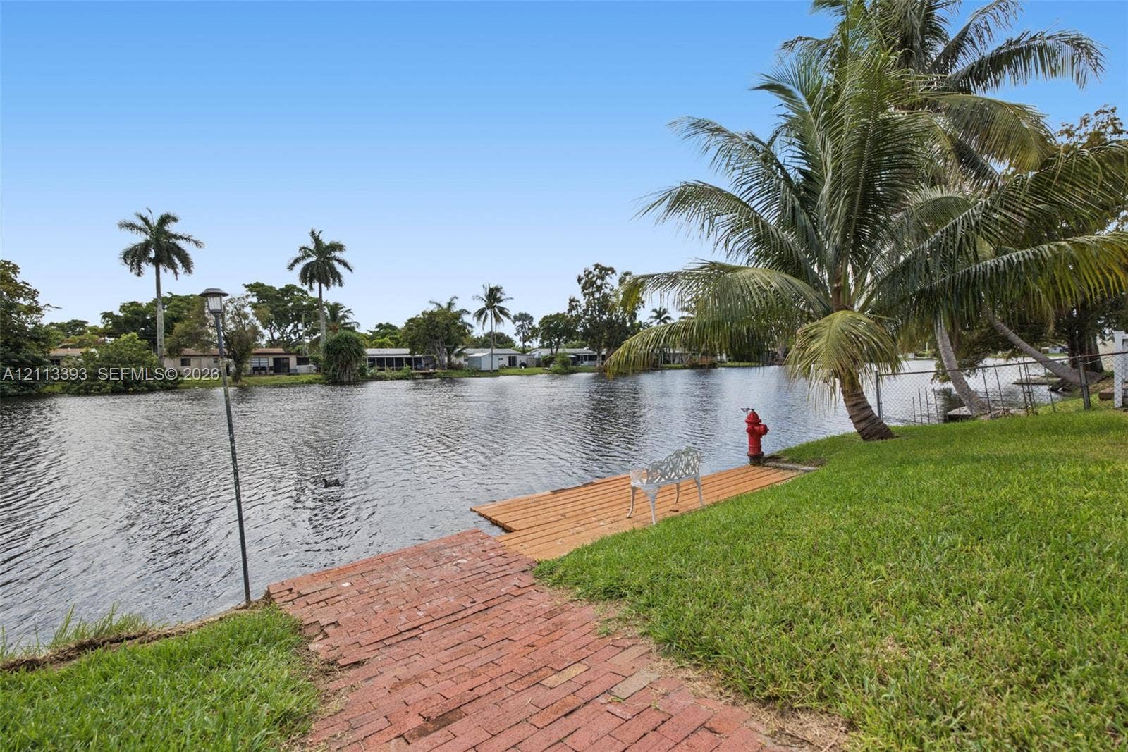 4136 SW 49th Street, Dania Beach, FL 33314 Waterfront property featuring a wood-plank dock, red brick path, manicured lawn, and mature palm trees