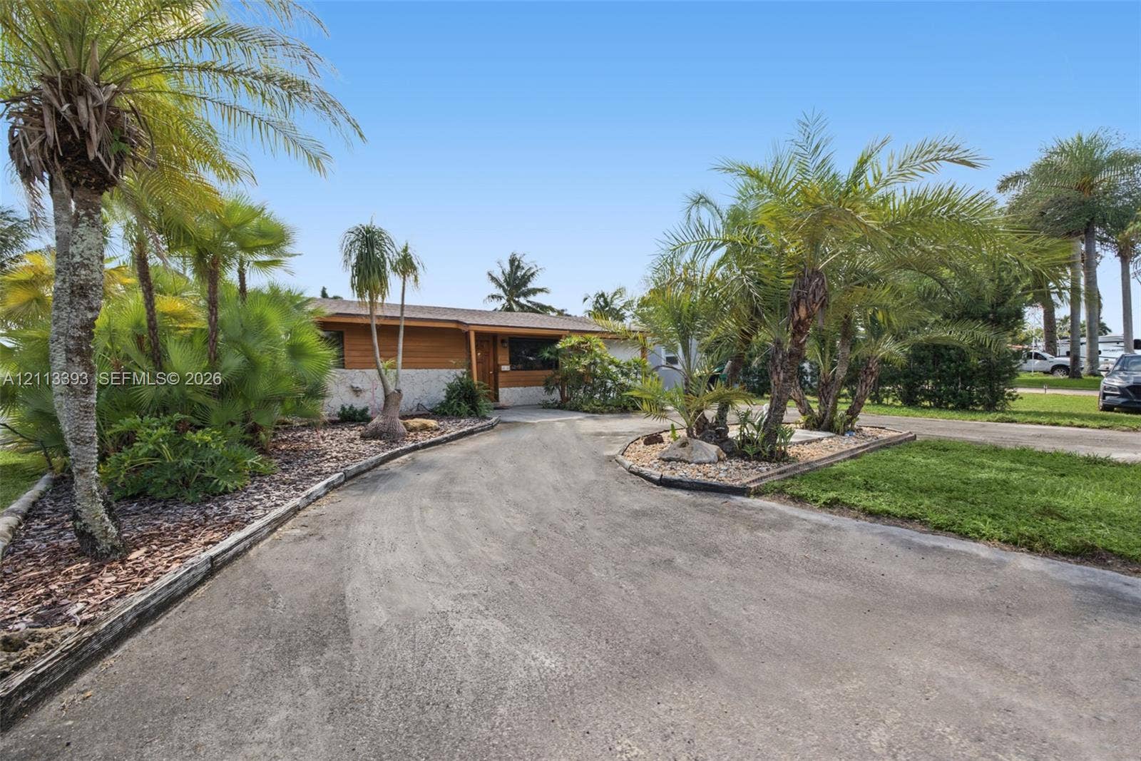 4136 SW 49th Street, Dania Beach, FL 33314 Exterior featuring horizontal wood siding, stone veneer accents, an expansive concrete driveway, and established palm tree landscaping