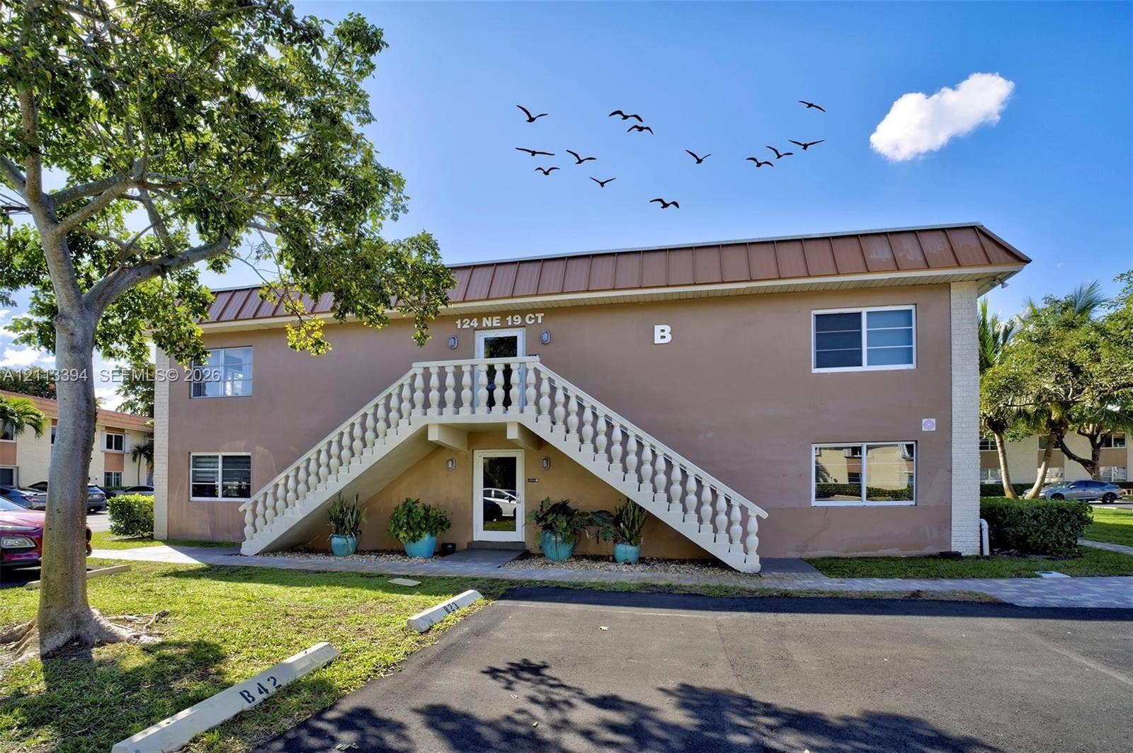 124 NE 19th Court, #108B, Wilton Manors, FL 33305 Two-story exterior featuring a metal roof, stucco finish, central staircase with a decorative balustrade, and paved parking