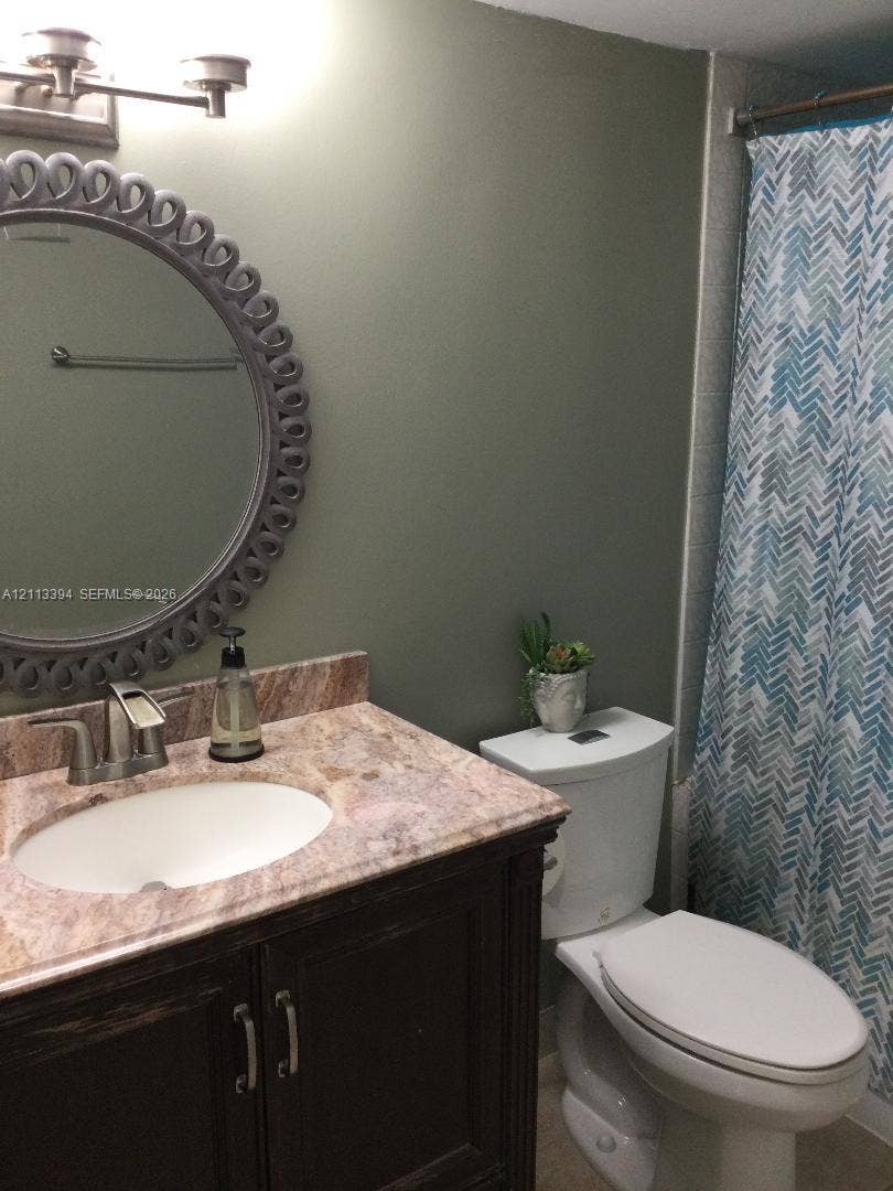 124 NE 19th Court, #108B, Wilton Manors, FL 33305 Bathroom featuring a dark wood-finish vanity, stone-patterned countertop, oval under-mount sink, brushed metal faucet, and decorative circular mirror