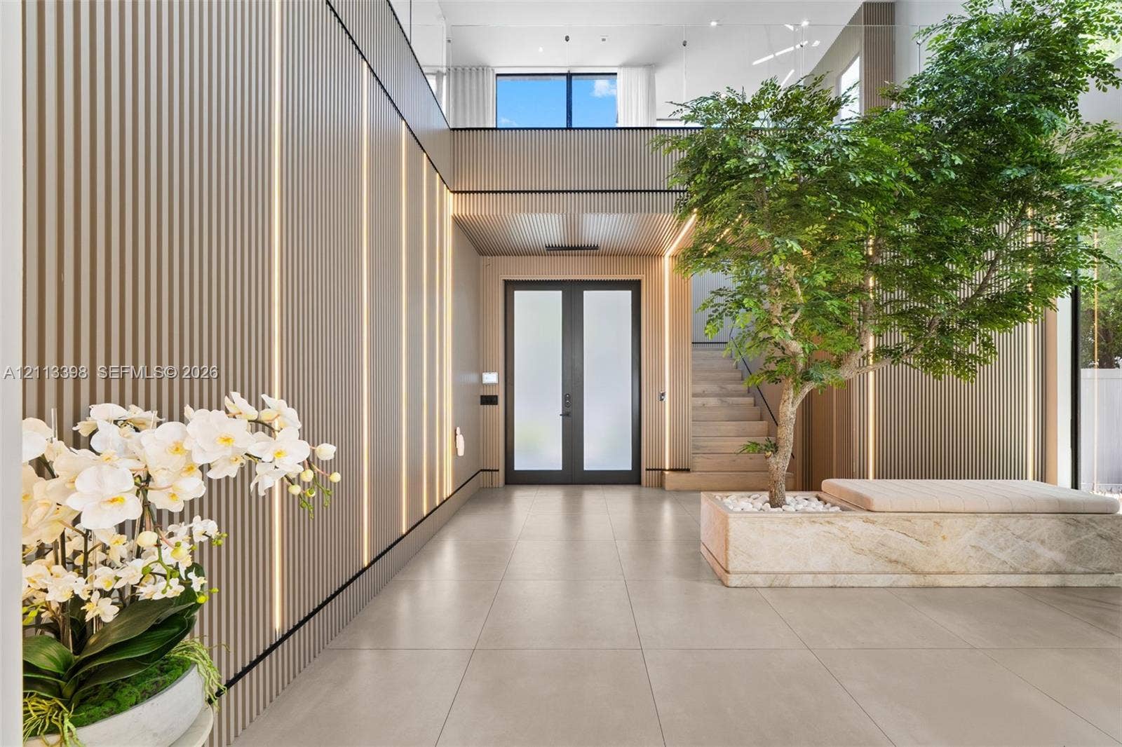 6814 NW 104th Court, #6814, Doral, Doral, FL 33178 Double-height foyer featuring vertical slat wall paneling with integrated recessed lighting, large-format tile flooring, a glass-railed staircase, and a stone-encased indoor planter